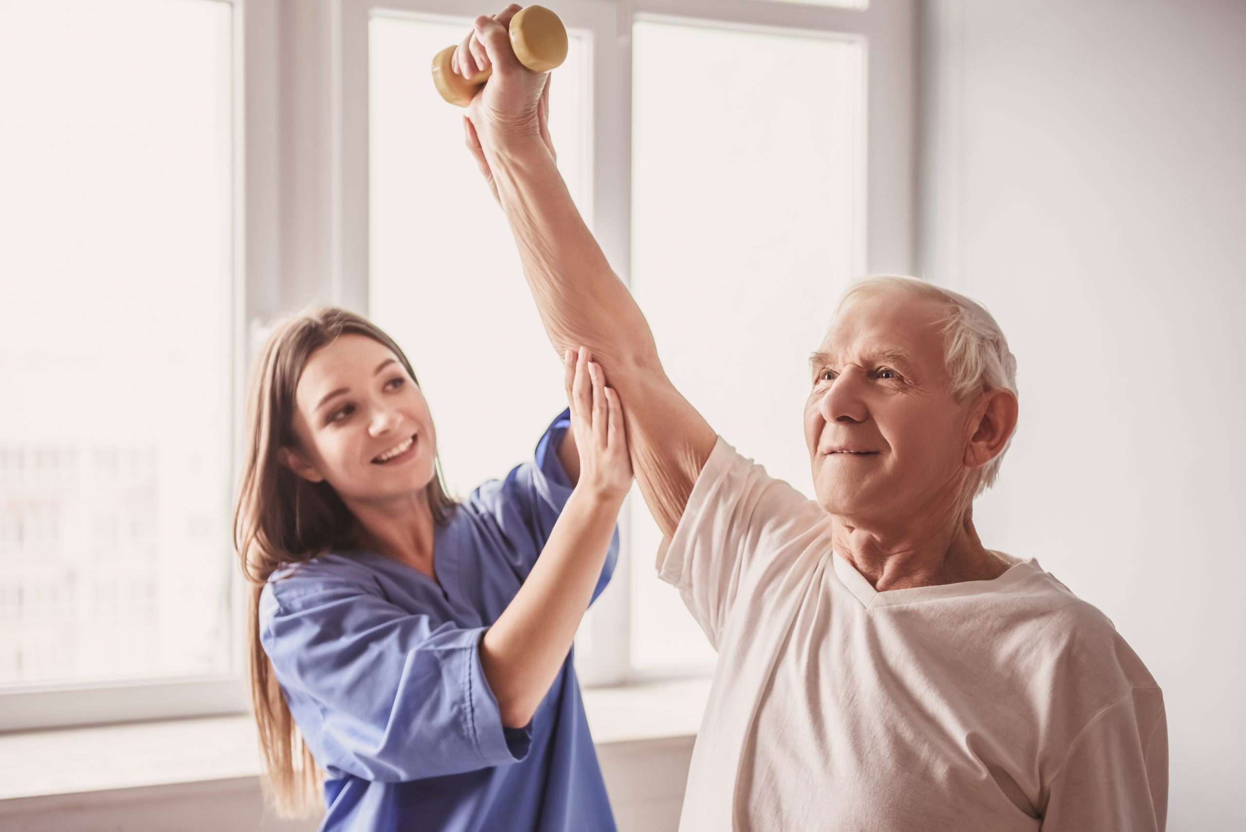Old man in hospital Handsome old man is doing exercises with dumbbells and smiling, in hospital ward. Attractive nurse is helping him